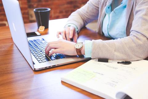 Woman typing on a laptop at a wooden table with a mug of tea beside here